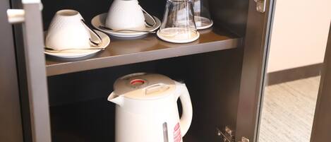 A kitchen area with a white electric kettle on the counter, a glass carafe, and white cups and saucers on the shelf above.