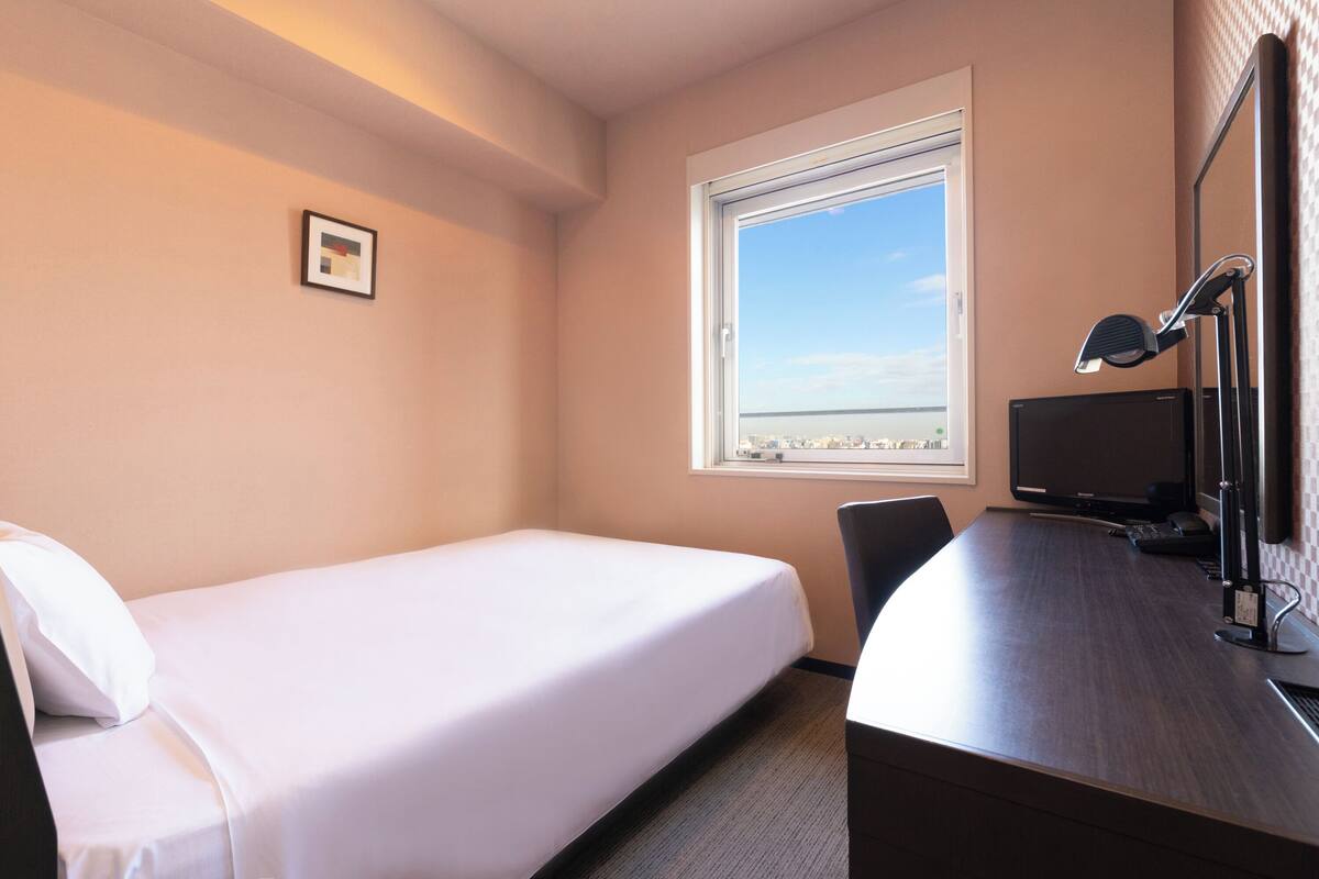 A hotel room with a single bed, a desk with a computer and a lamp, a window with a view of the city, and a framed picture on the wall.