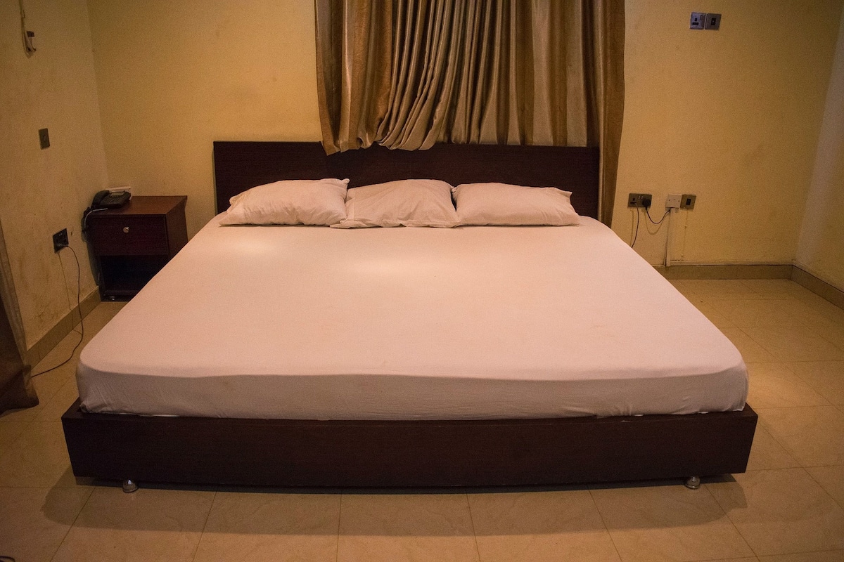 A neatly made bed with a dark headboard, a bedside table with a phone, and a power outlet on the wall.