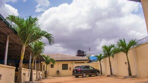 A paved courtyard with a parked SUV, palm trees, and a building with a tiled roof. - Taverna Guest House (Lagos)