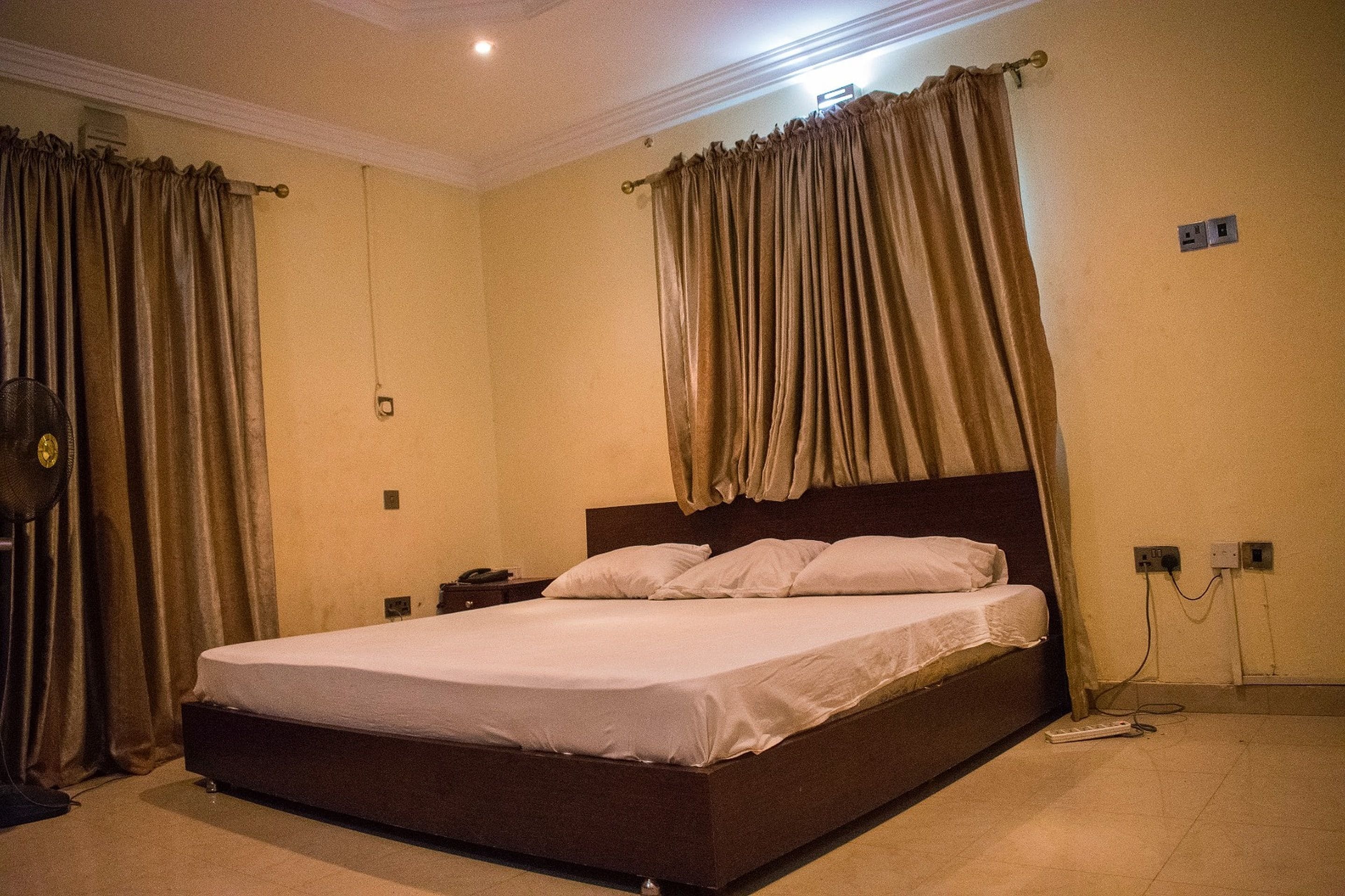 Quarto de hotel padrão com uma cama de solteiro, mesa de cabeceira, ventoinha e cortinas.
