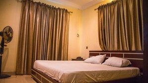 A neatly made bed with a wooden headboard, a bedside table with a lamp, and curtains on the windows. - Taverna Guest House (Lagos)