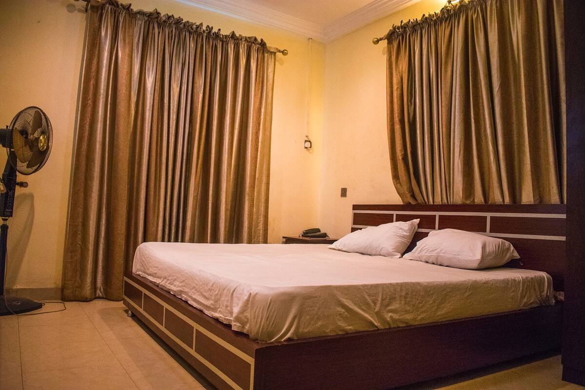 A neatly made bed with a wooden headboard, a bedside table with a lamp, and curtains on the windows.