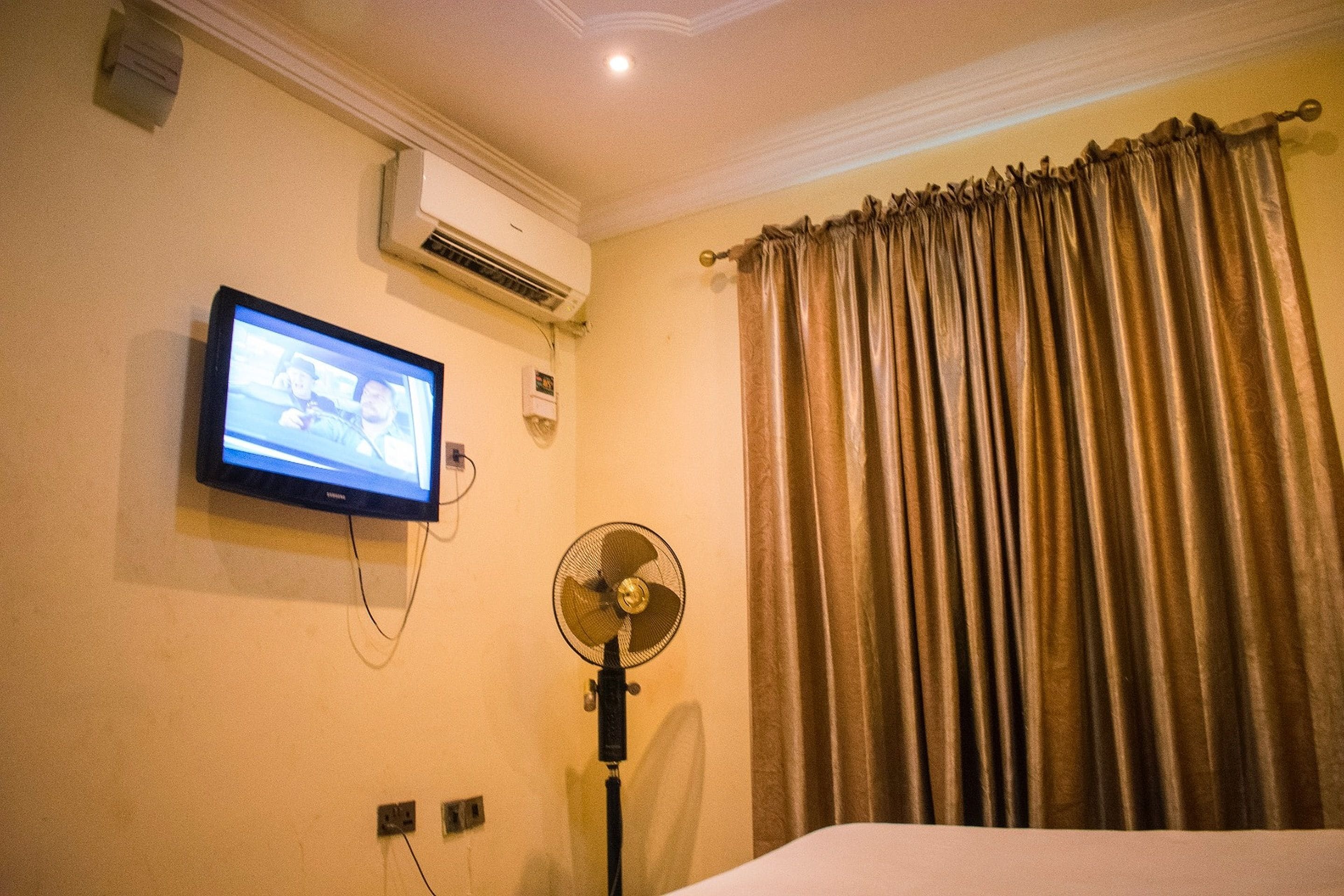 A hotel room with a bed, an air conditioning unit, a television mounted on the wall, and a standing fan.
