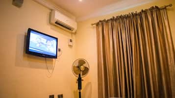 A hotel room with a bed, an air conditioning unit, a television mounted on the wall, and a standing fan.