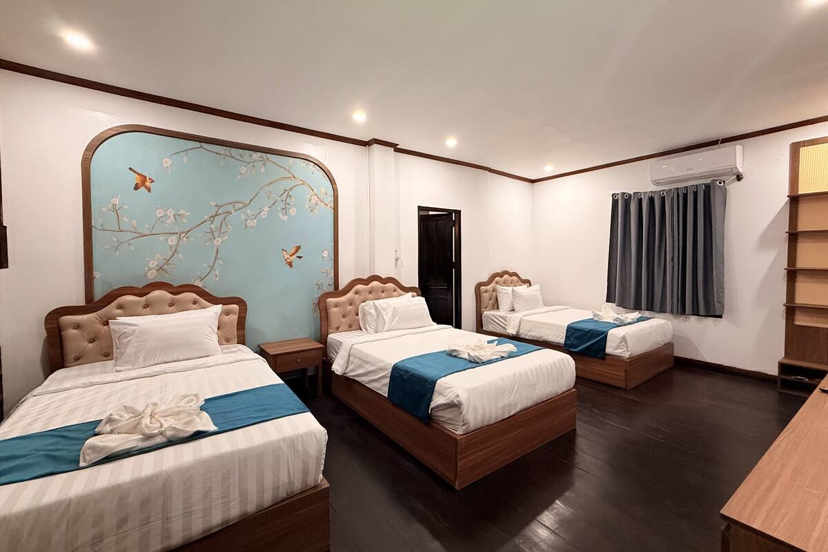 Une chambre d’hôtel avec trois lits, une grande fenêtre avec des rideaux, un plancher en bois et un mur orné d’une fresque florale.