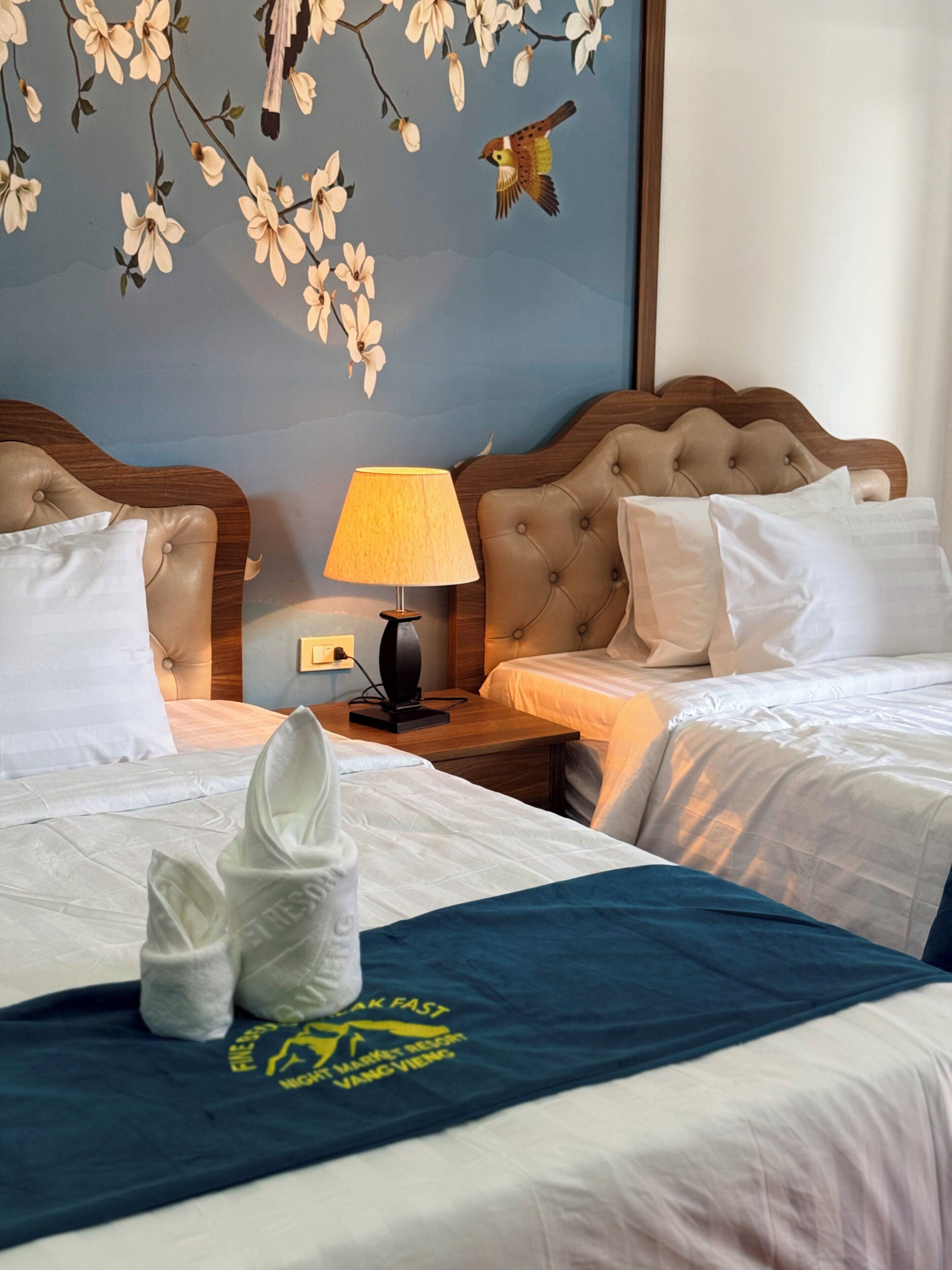 A hotel room with two beds, a nightstand with a lamp, and a wall with floral wallpaper.