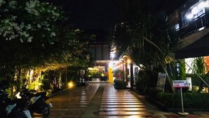 Une vue nocturne de l’entrée d’un bâtiment, avec un comptoir d’accueil, un chemin et des motos garées.