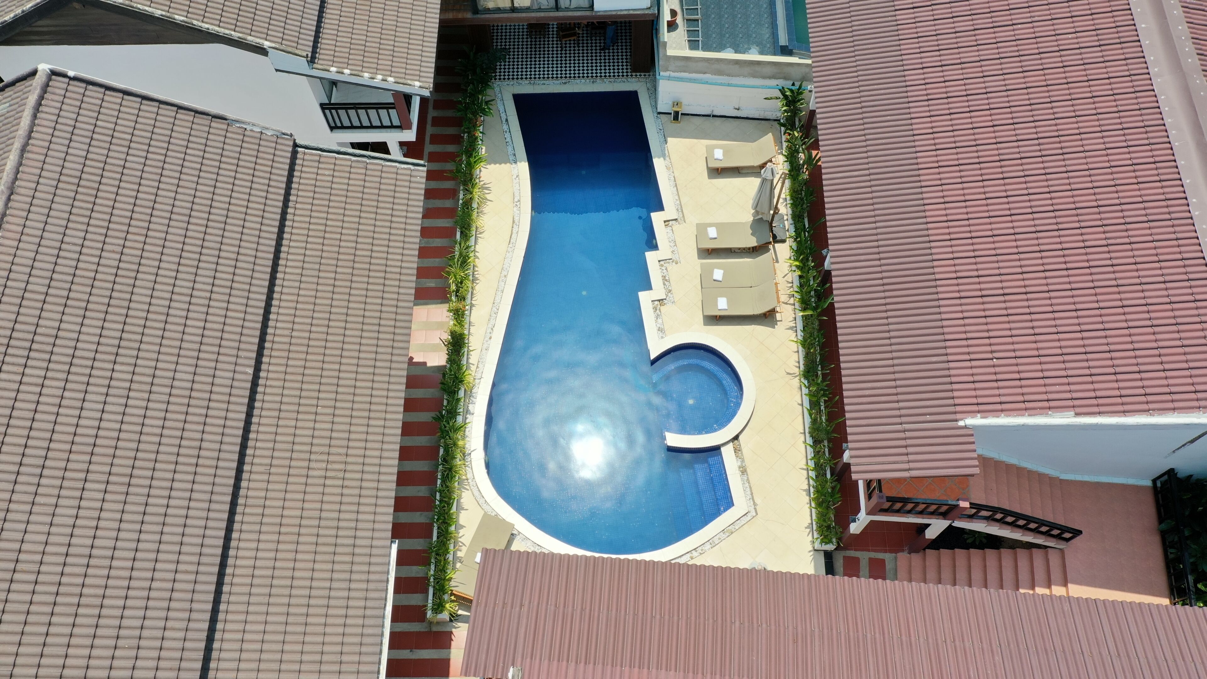An aerial view of a swimming pool with a small circular section, surrounded by a tiled deck and a few lounge chairs.