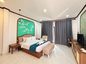 Un dormitorio con cama, mesitas de noche, un banco, un escritorio con silla, televisor y un amplio ventanal con cortinas.
