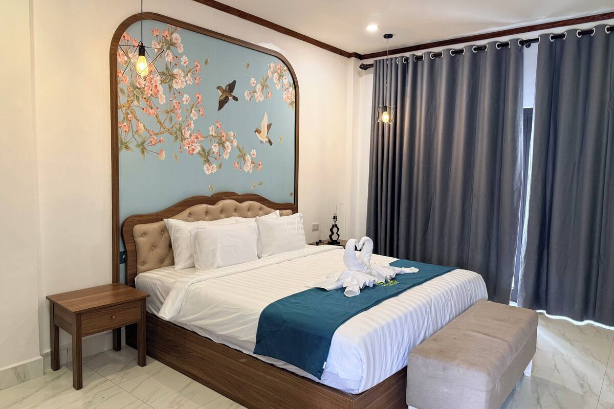 Une chambre à coucher comprenant un lit, une table de chevet, un banc, un mur décoré de motifs floraux et d’oiseaux, et une fenêtre avec des rideaux.