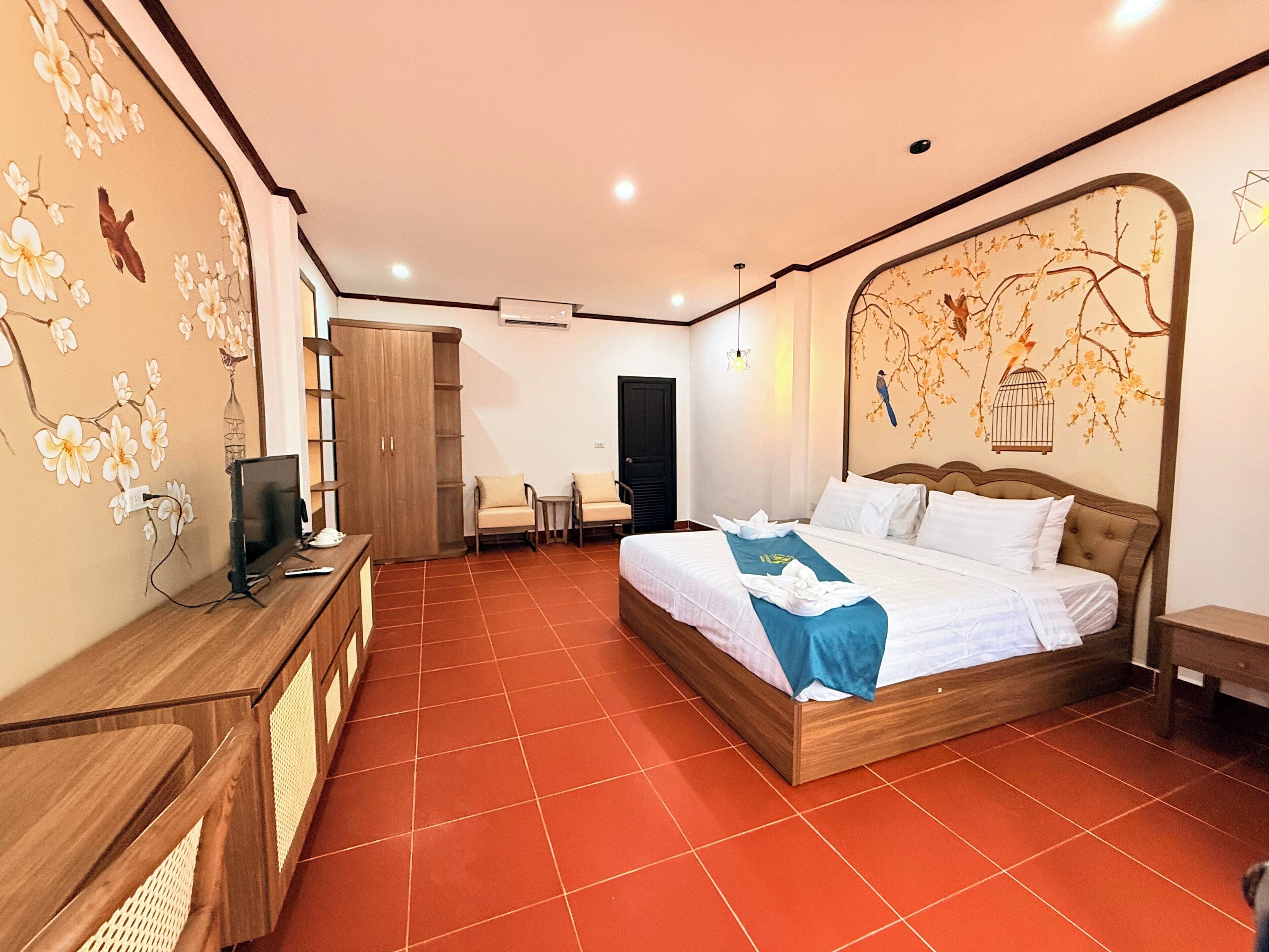 A hotel room with a large bed, a desk with a computer, a wardrobe, and a wall with floral and bird artwork.