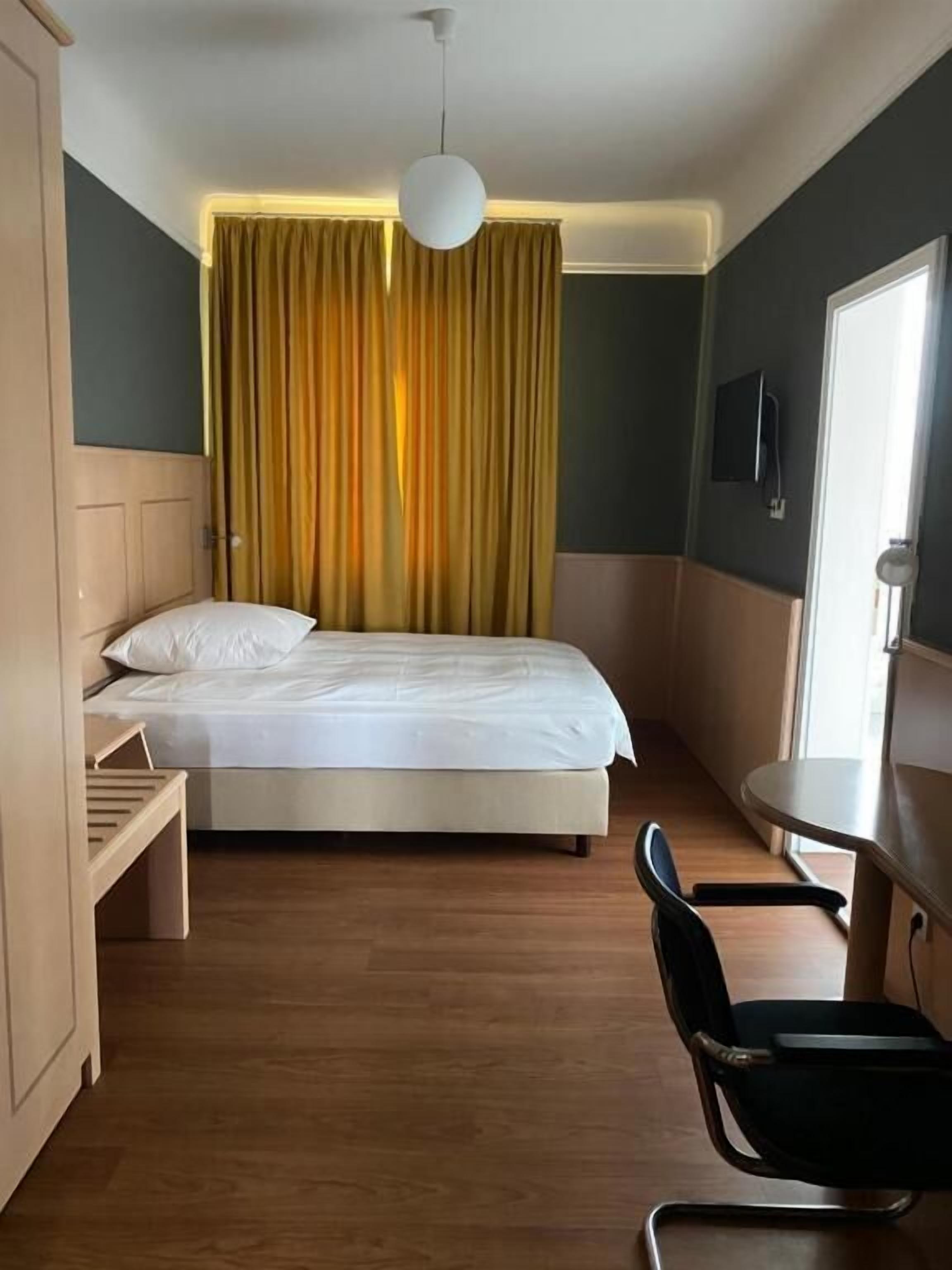 Habitación de hotel con cama, escritorio, silla, televisor y ventana con cortinas.