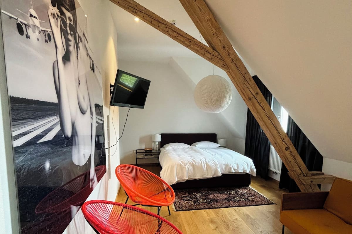 A bedroom with a sloped ceiling, a bed, a TV, and two red chairs.