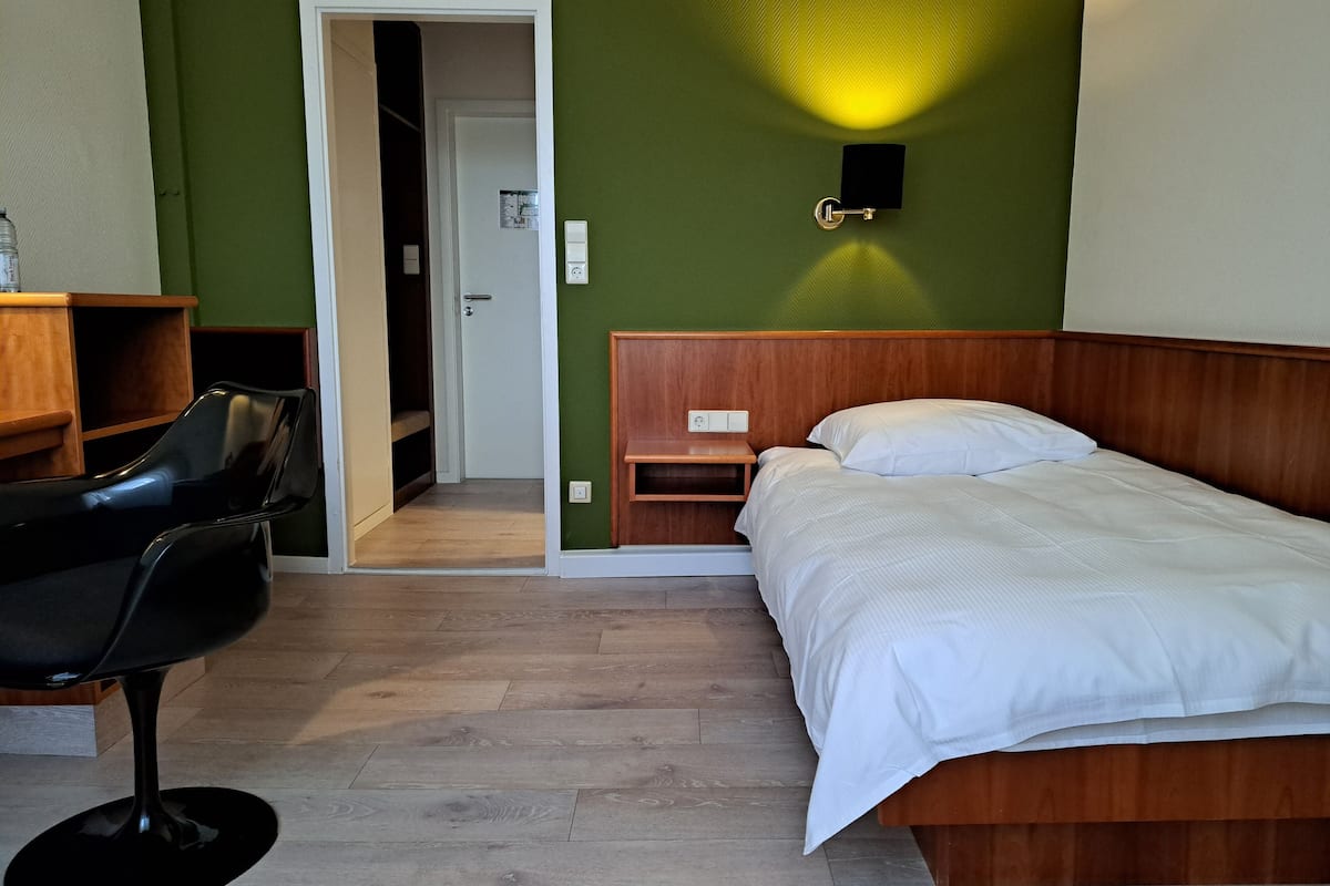 A room with a bed, a chair, a wooden nightstand, and a green wall with a wall-mounted light fixture.