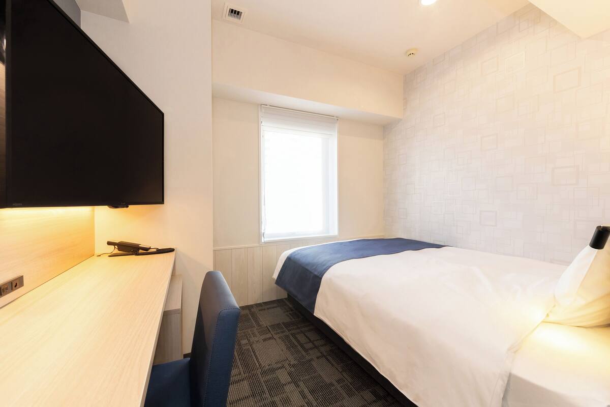 A hotel room with a bed, a desk, a chair, a window, and a flat-screen TV.