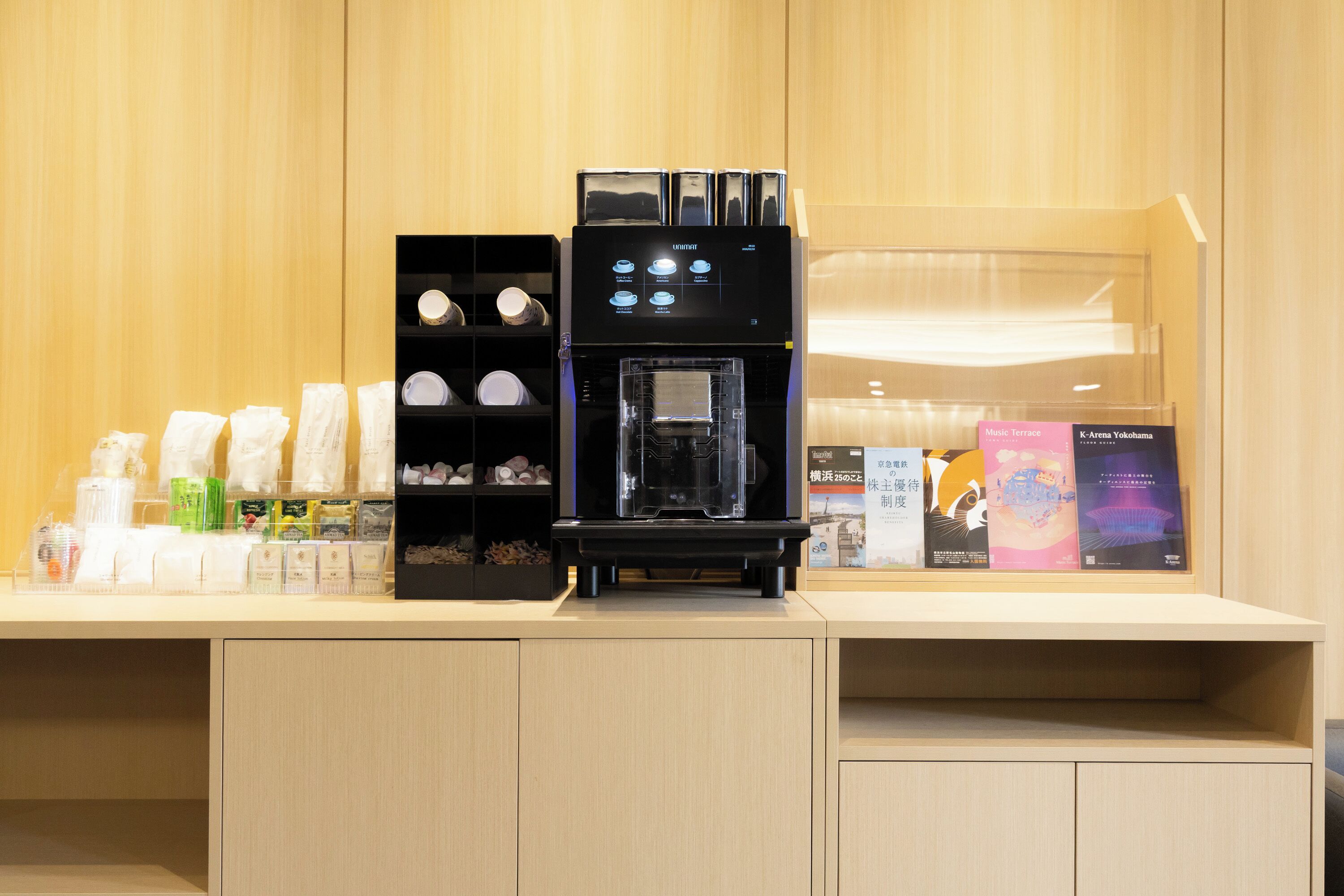 A coffee machine in a hotel lobby with a wooden panel background and a display of books and magazines.