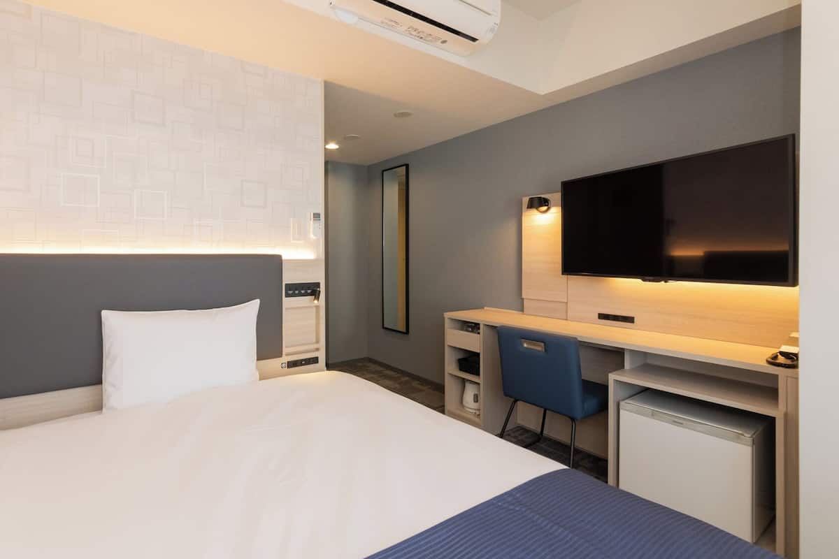 Quarto de hotel com cama, escrivaninha, cadeira, televisão e espelho.