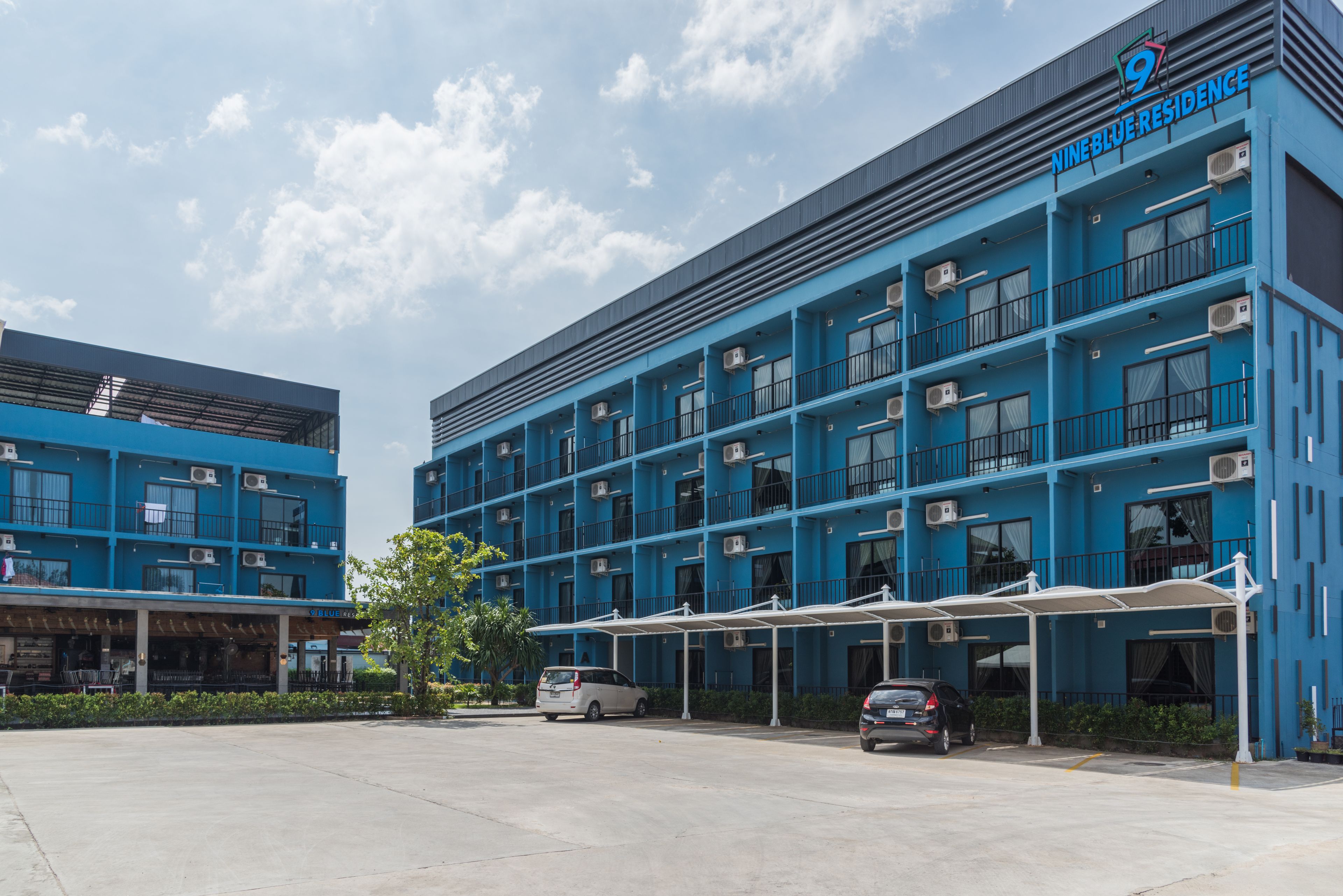 Um hotel com vários andares, fachada azul, unidades de ar condicionado e área de estacionamento coberta.