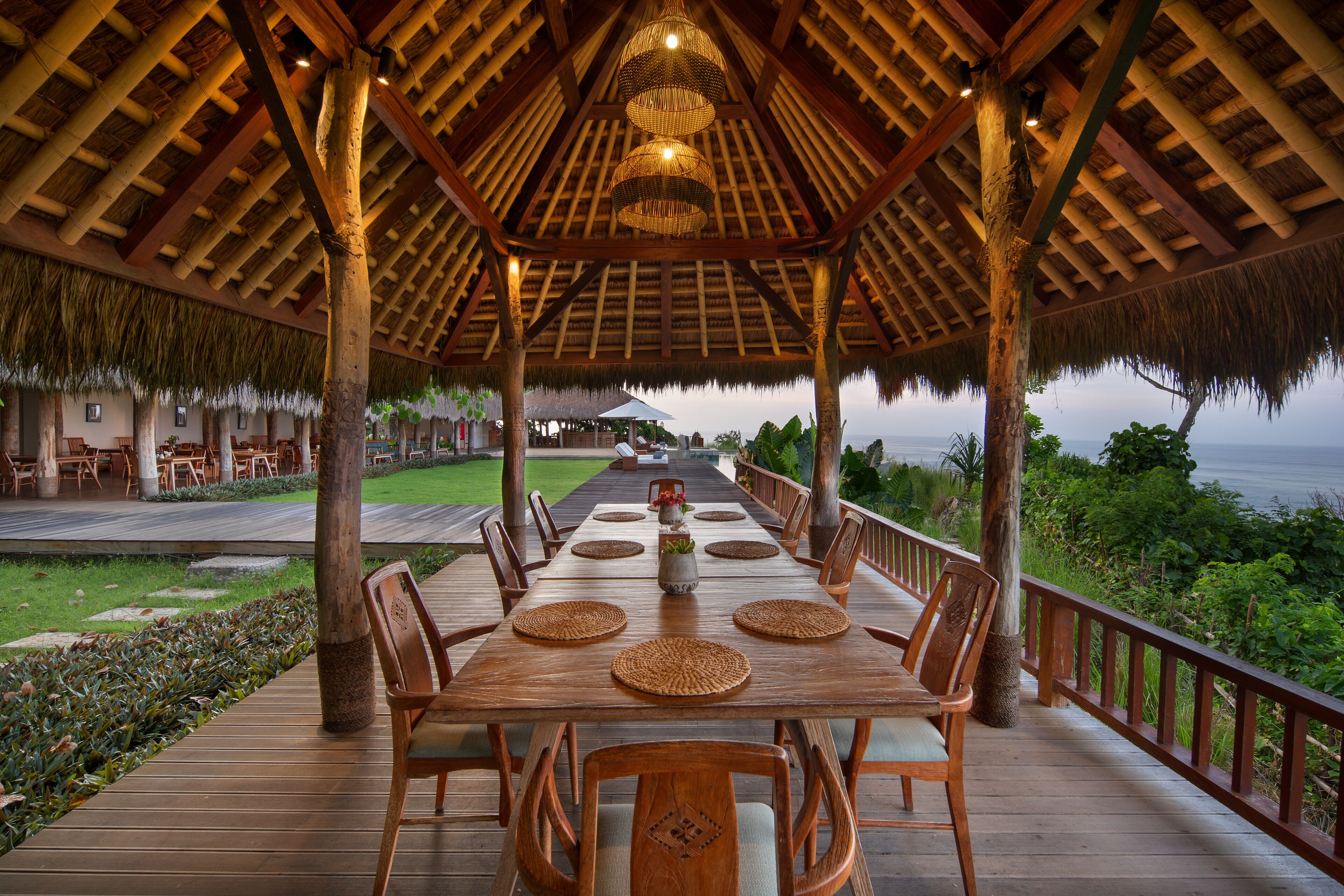 Una terraza de madera con techo de paja, una larga mesa dispuesta para comer, y vista al océano.