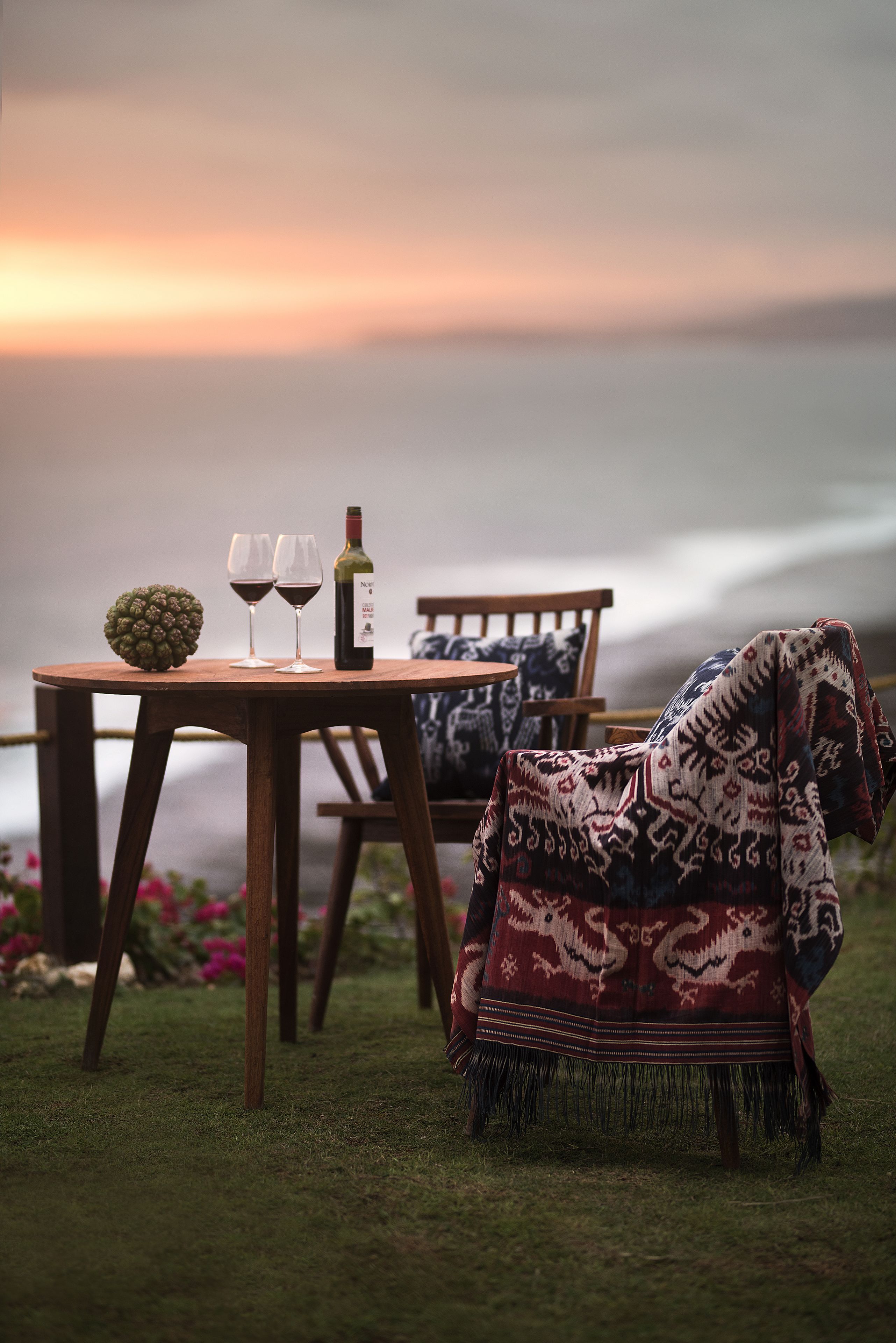 A table with two wine glasses and a bottle, a fruit, and a patterned blanket on a grassy area near a body of water.