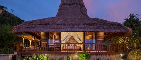 A thatched-roof bungalow with a balcony, surrounded by lush tropical vegetation.