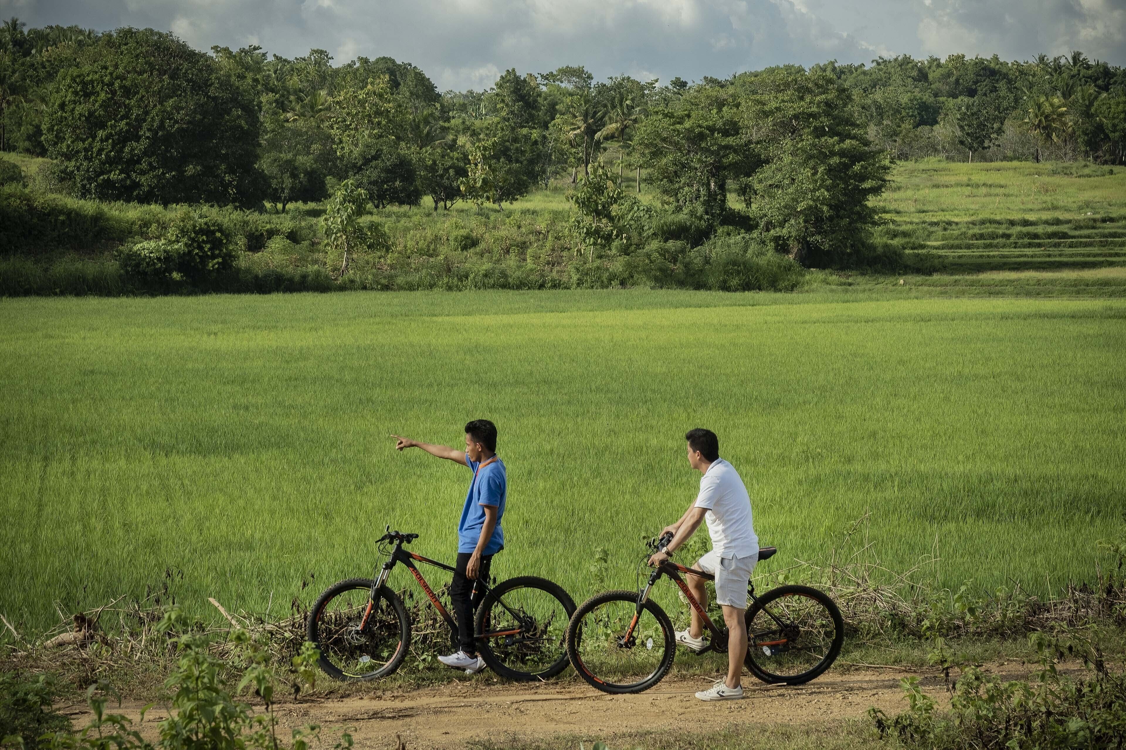 Deux personnes à vélo dans un champ verdoyant, avec des arbres en arrière-plan.
