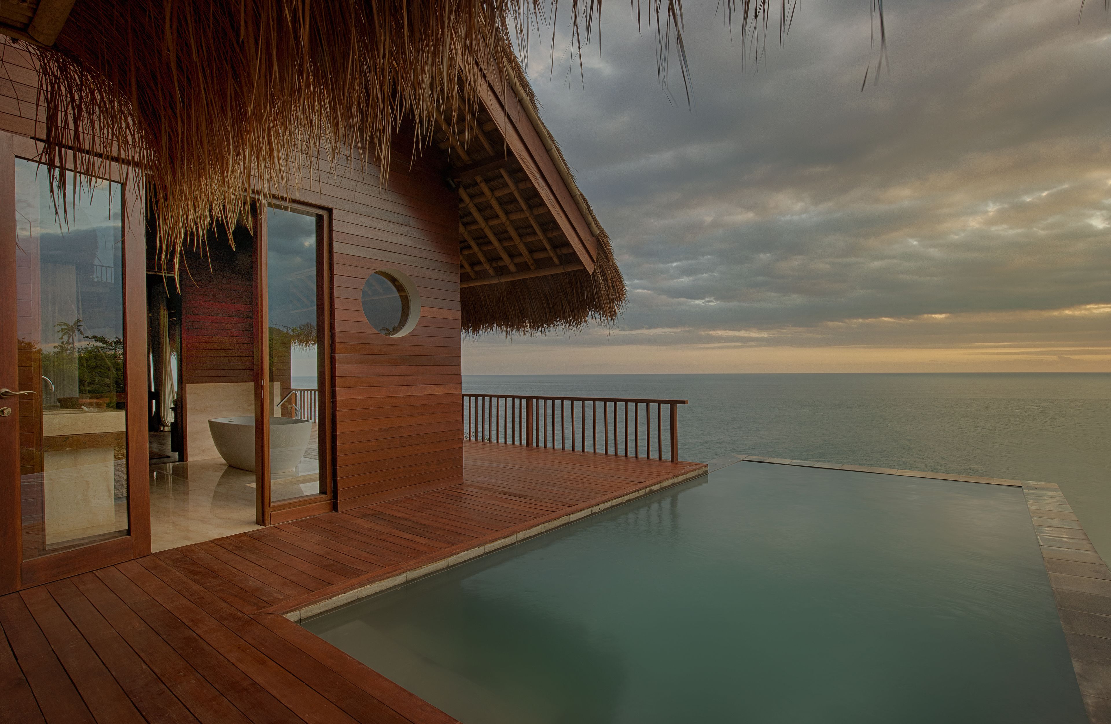 A wooden infinity pool with a glass balustrade, a thatched-roof structure, and a view of the sea.