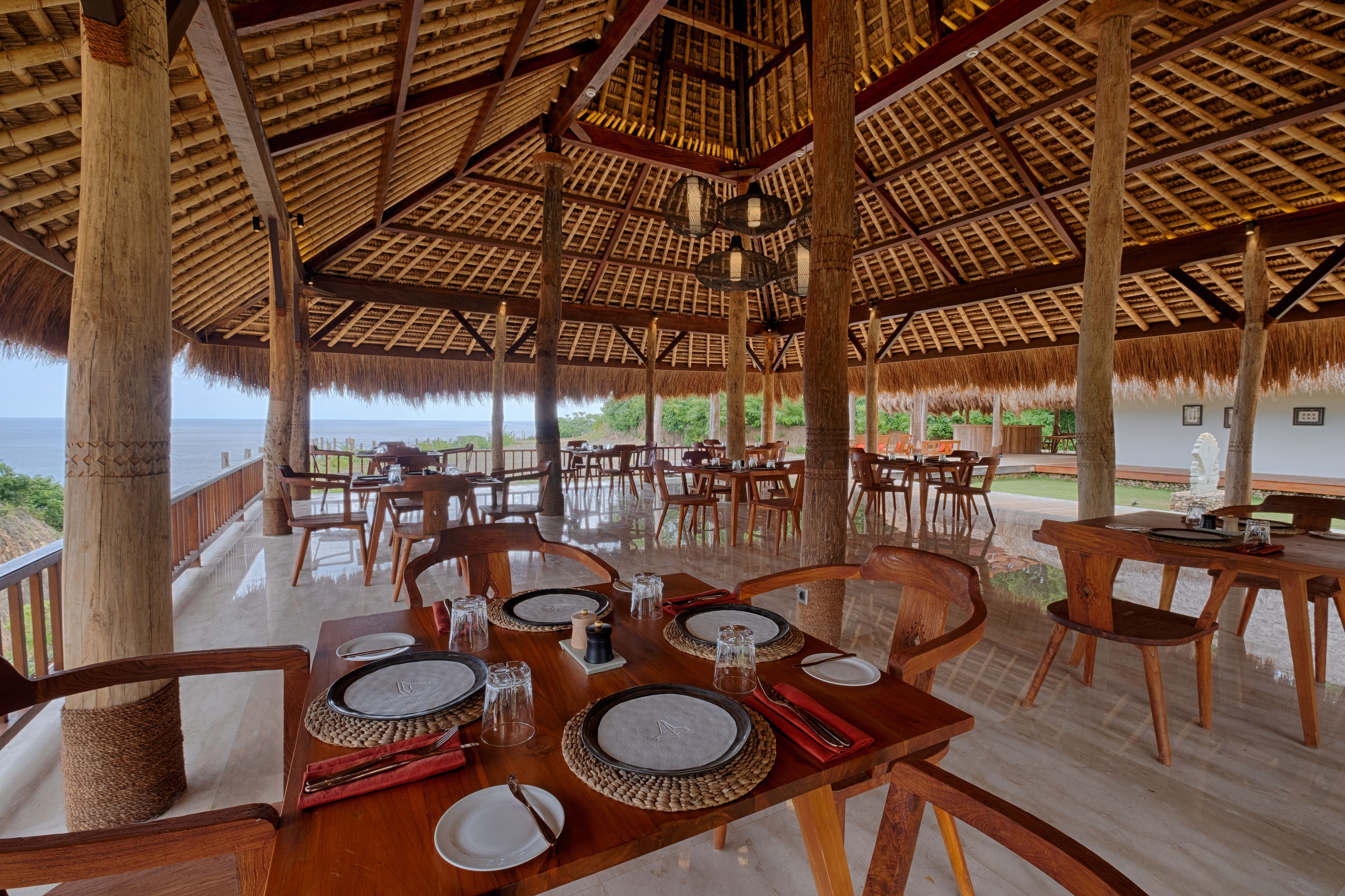 An outdoor dining area with wooden tables and chairs, thatched roof, and a view of the ocean.
