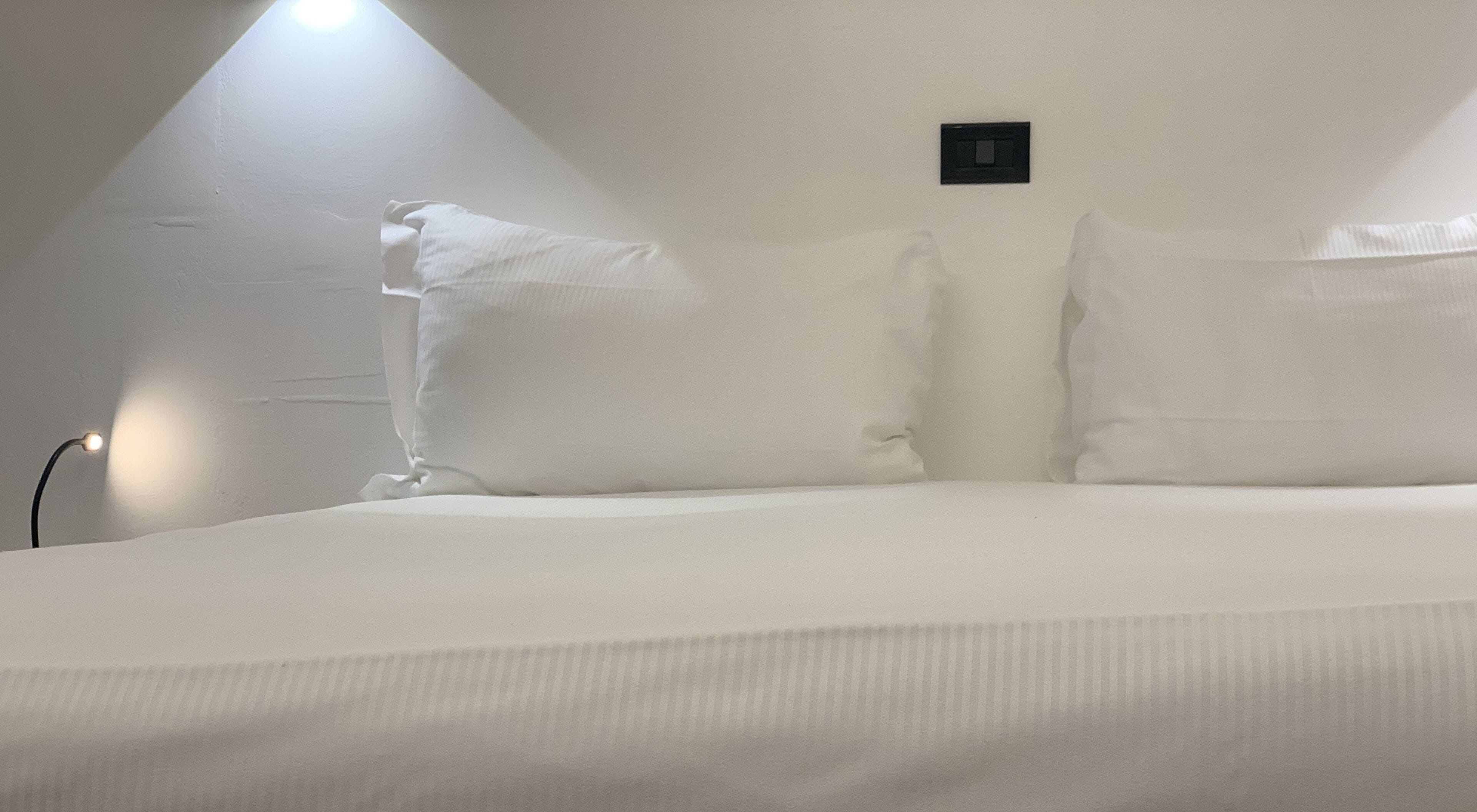 Un dormitorio con una cama individual, ropa de cama blanca y una lámpara de pared pequeña.