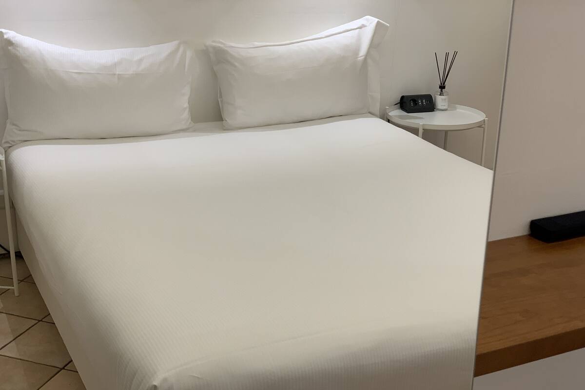 A neatly made bed with white bedding, a small bedside table with a radio and a bottle of reed diffuser, and a wooden shelf.