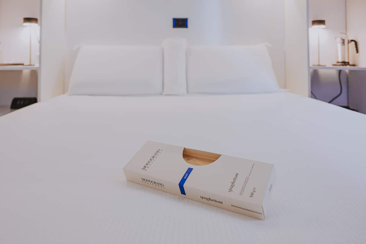 A neatly made bed with a box of Monograno Spaghetti on it.