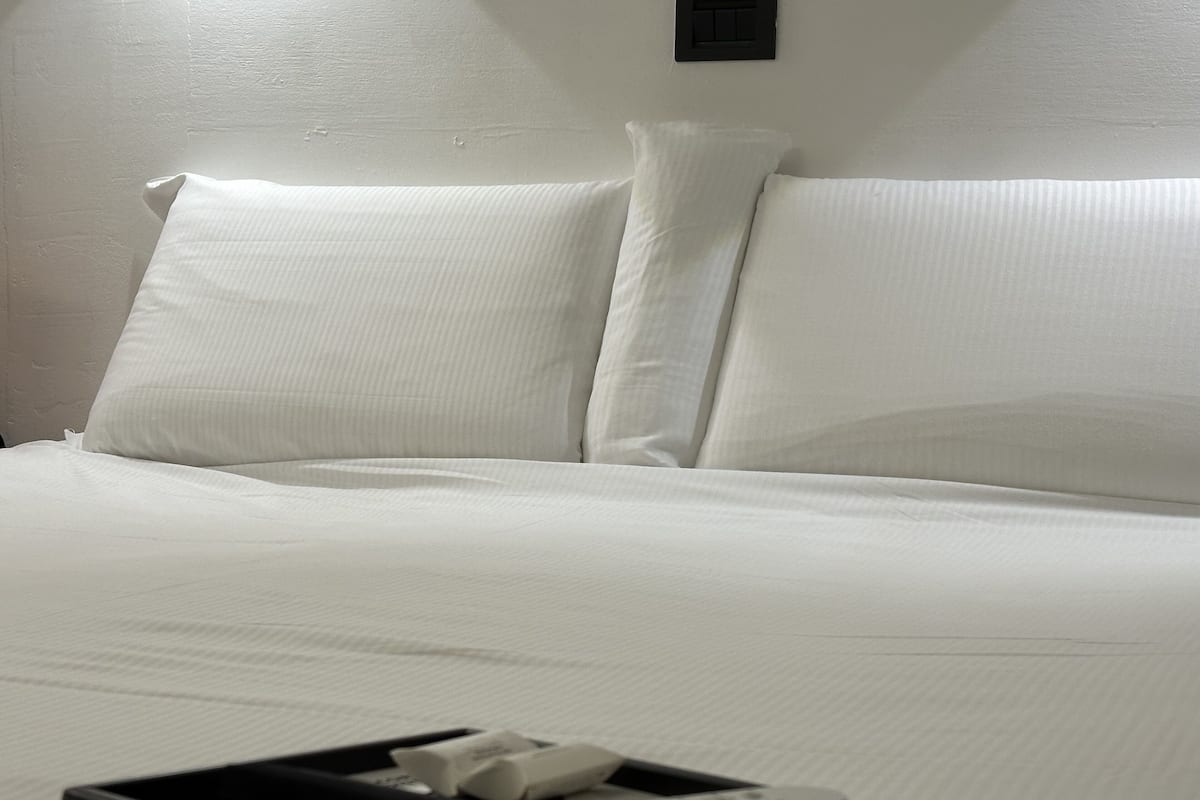 A neatly made bed with white linens and pillows, a black tray with a remote control and a book, and a wall switch above the bed.