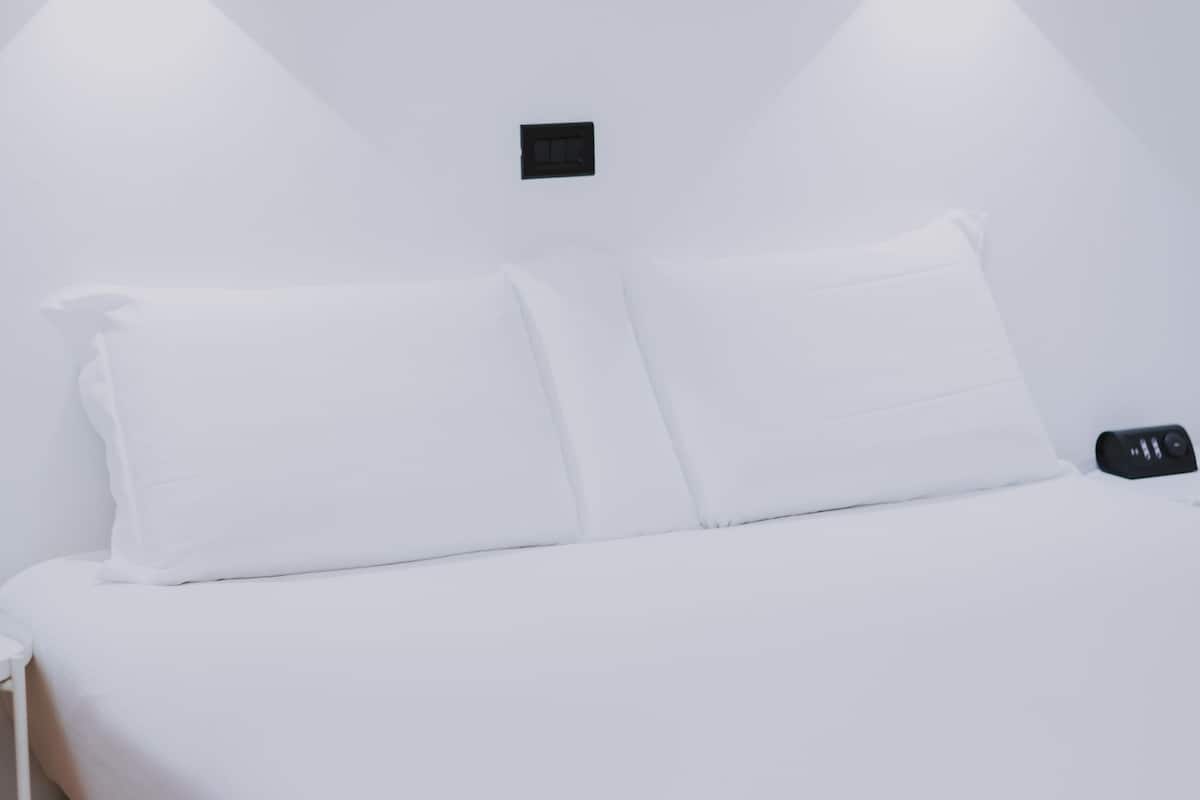 A minimalist hotel room with a neatly made bed, a bedside table, and a wall-mounted light fixture.