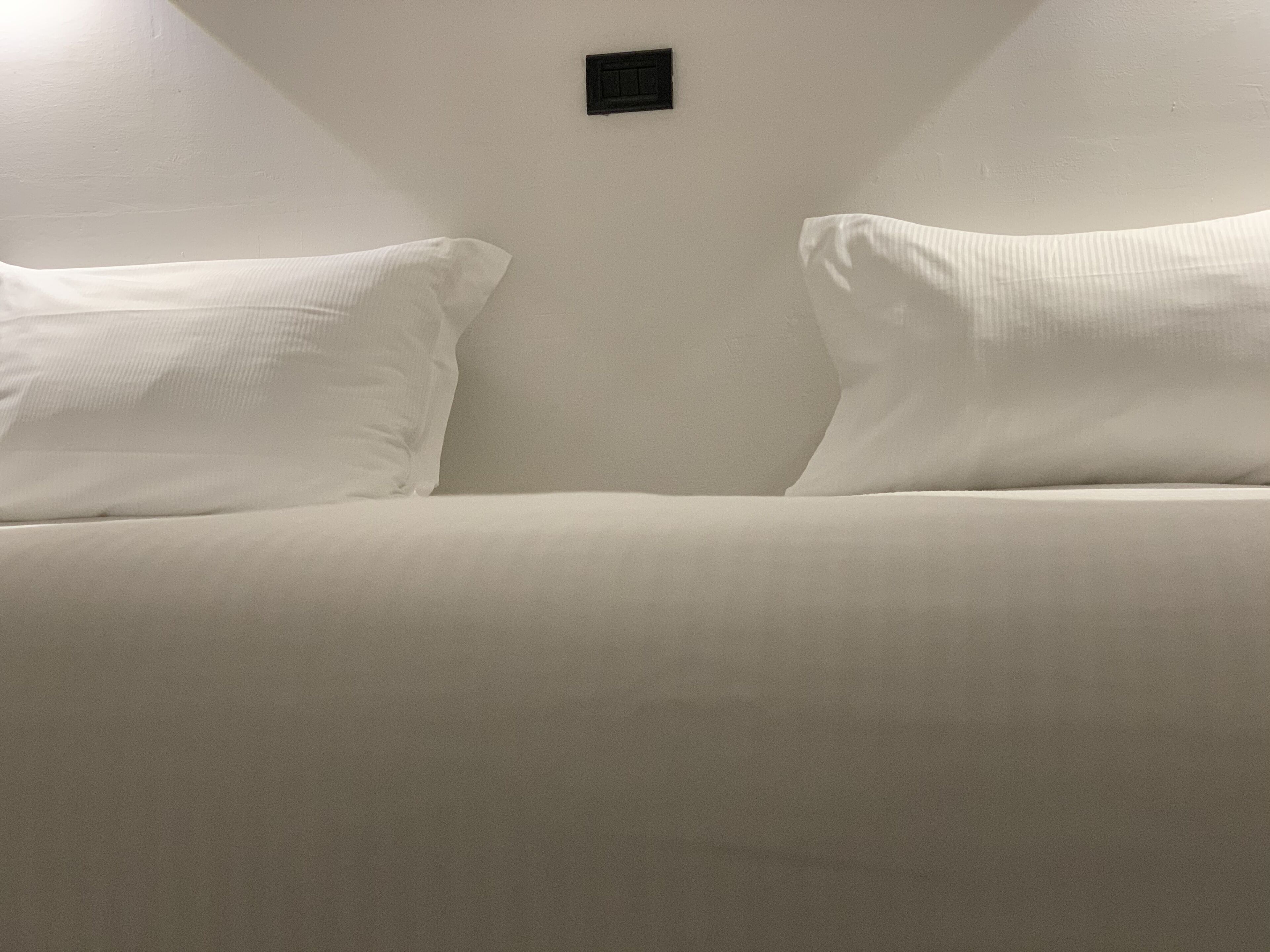 Sängyssä on kaksi tyynyä ja seinällä pieni musta esine.