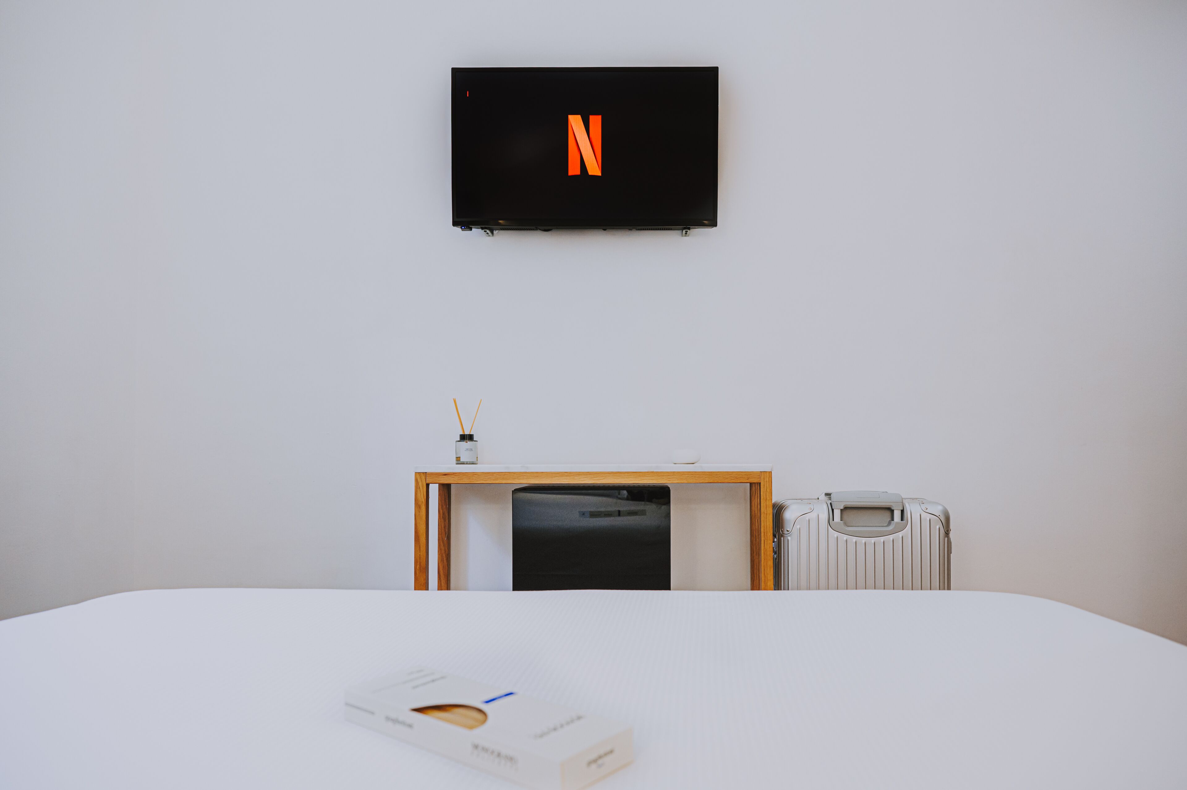A hotel room with a bed, a small table with a box, a radiator, and a TV mounted on the wall displaying a Netflix logo.