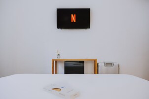 A hotel room with a bed, a small table with a box, a radiator, and a TV mounted on the wall displaying a Netflix logo.