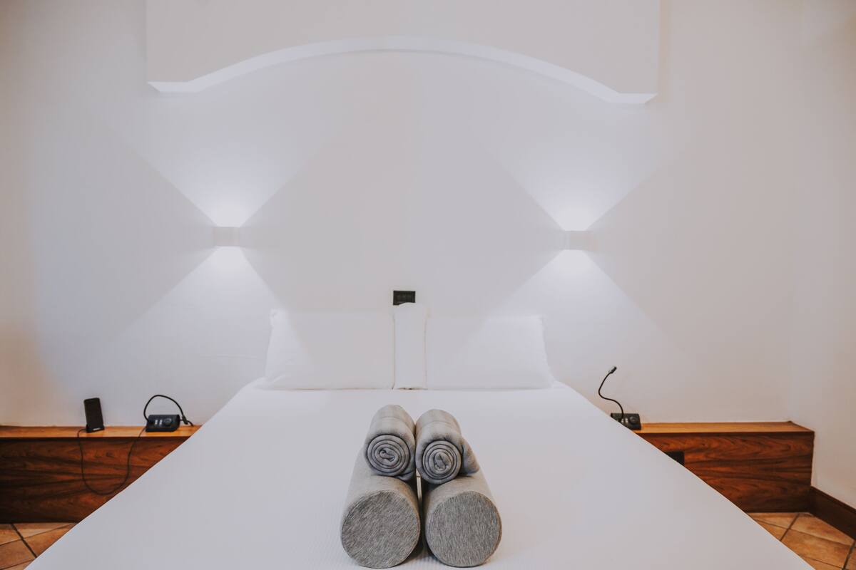 A neatly made bed with rolled towels, a bedside table with a speaker and phone, and a wooden bench along the wall.