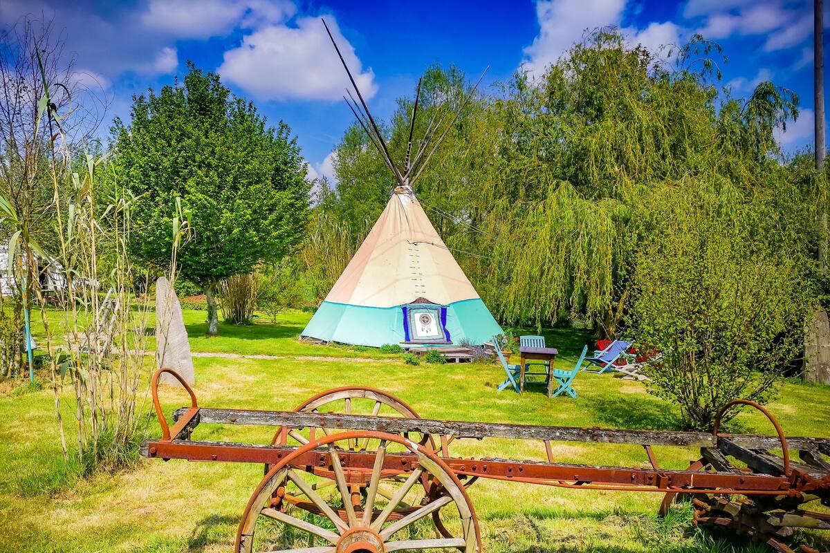 A traditional tipi with a blue and beige exterior, surrounded by a grassy area with trees and a wooden cart.