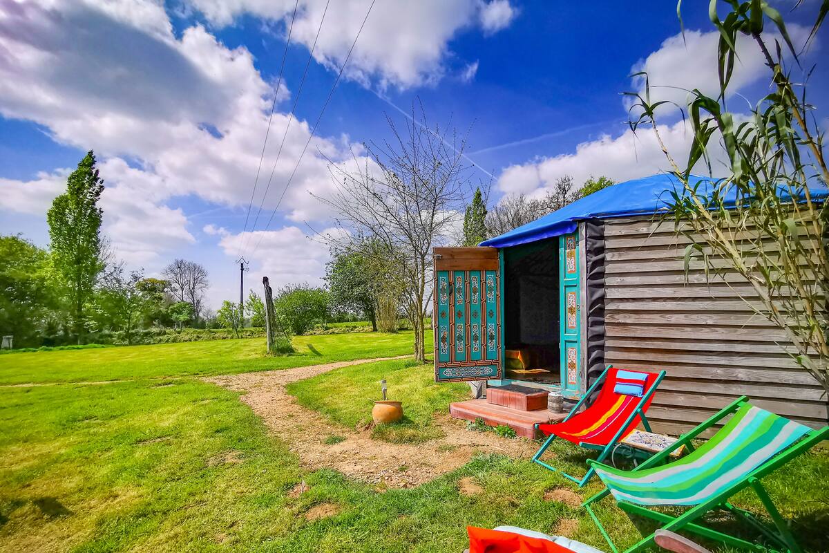A wooden cabin with a blue roof, a garden with a dirt path, and colorful beach chairs.