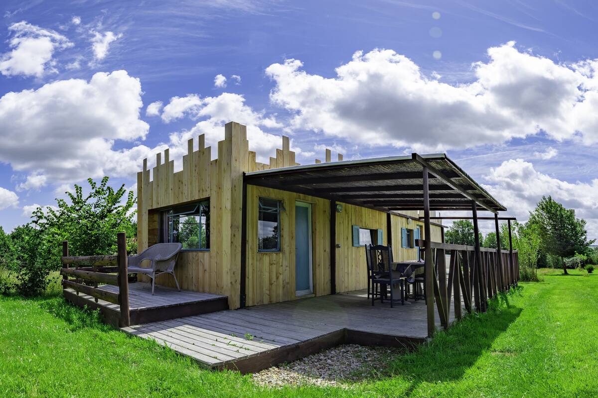 A wooden cabin with a deck, a covered patio, and a view of a lush green landscape.