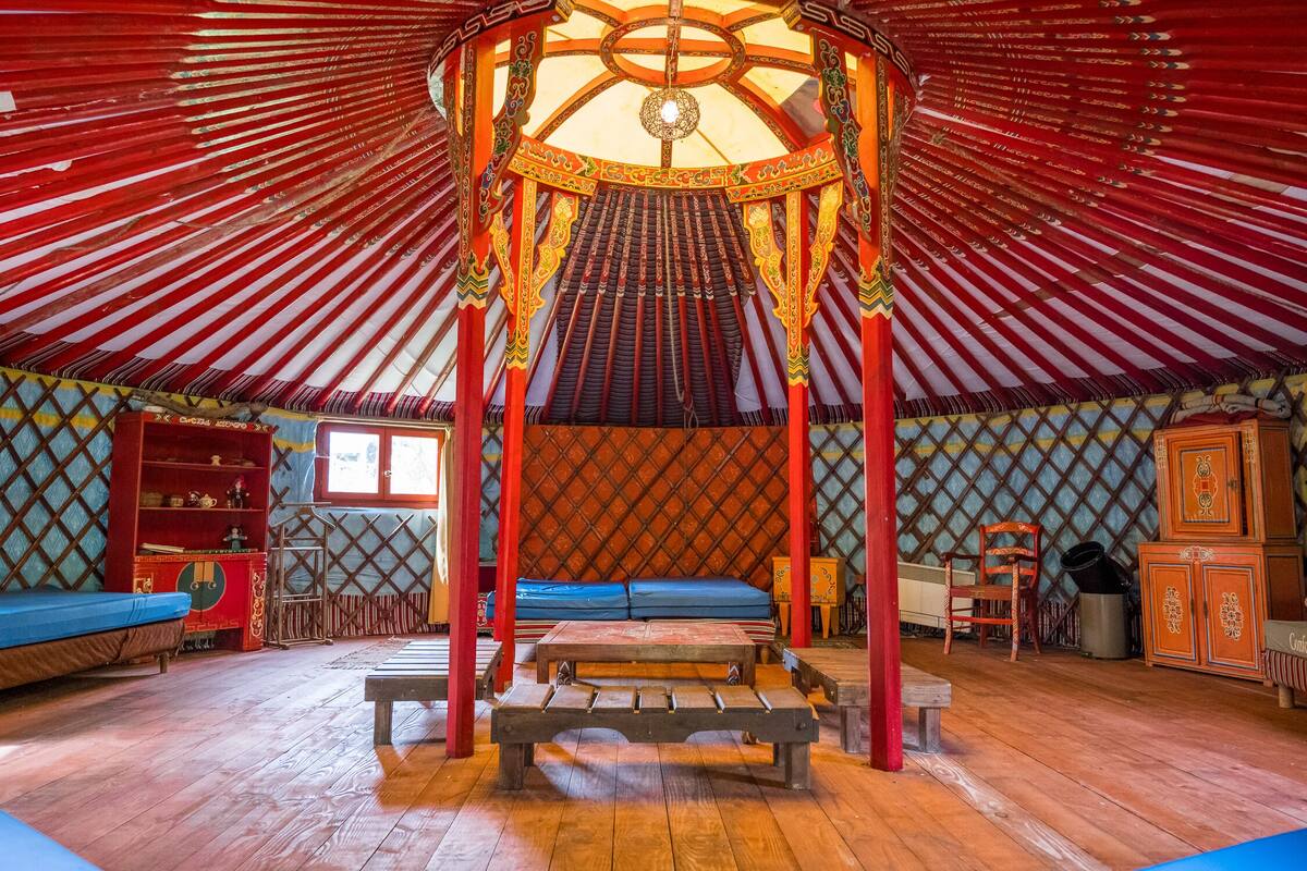 A traditional yurt interior with a central bed, wooden benches, and a decorative ceiling.
