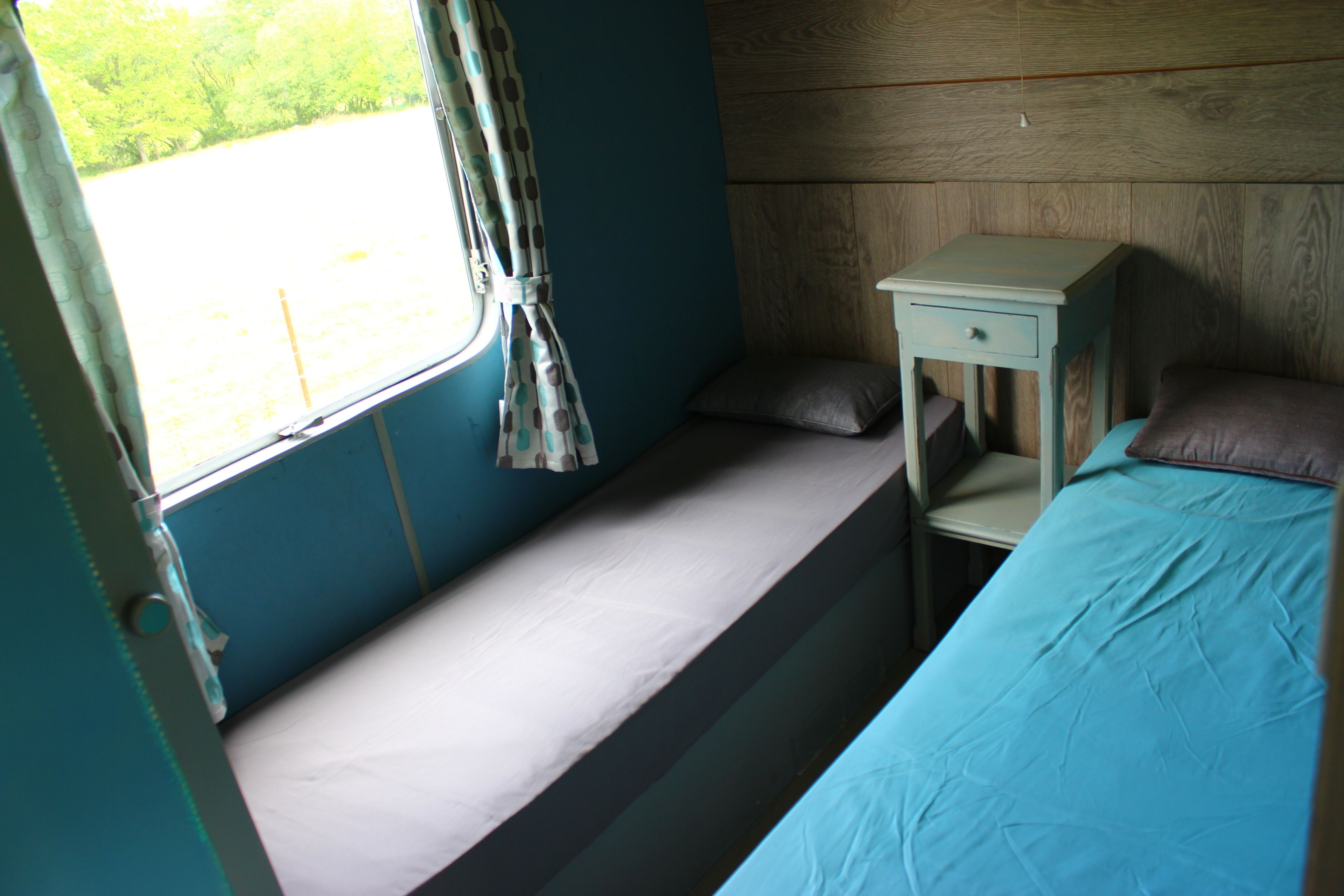 A small, single-bed room with a window, a bedside table, and a pillow.