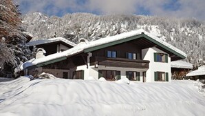 Un grand chalet à plusieurs étages, doté d’un toit en pente, entouré d’arbres et de montagnes couverts de neige.