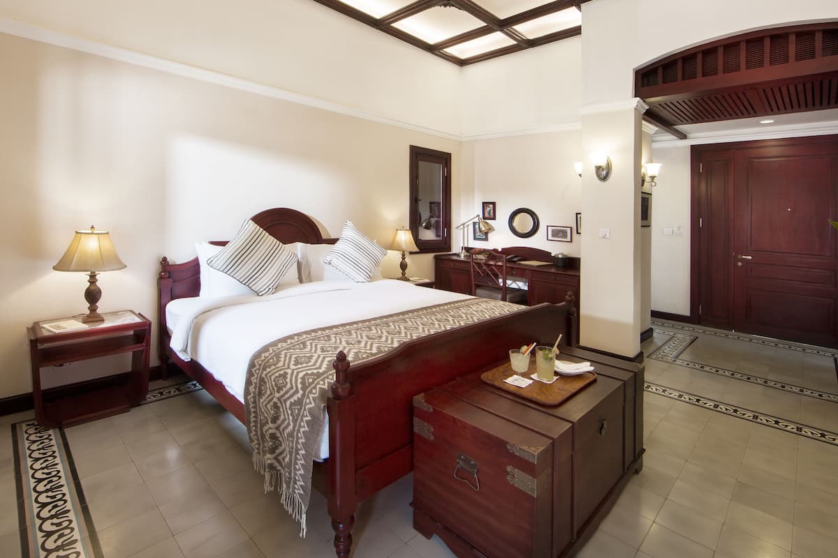 A hotel room with a large bed, a nightstand with a lamp, a dresser, and a wooden door.