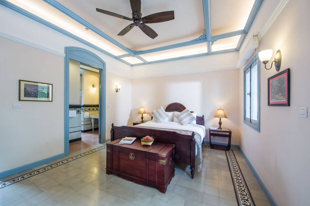 A bedroom with a bed, bedside lamps, a ceiling fan, a wooden chest of drawers, and a patterned floor.