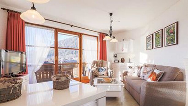 A cozy living room with a sofa, coffee table, and a TV. There is a window with curtains and a view of the outdoors. - Ferienwohnung Lea Stübchen (Reit im Winkl)
