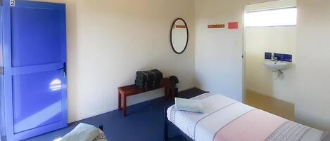 A room with a bed, a bench, a blue door, a mirror, and a sink.