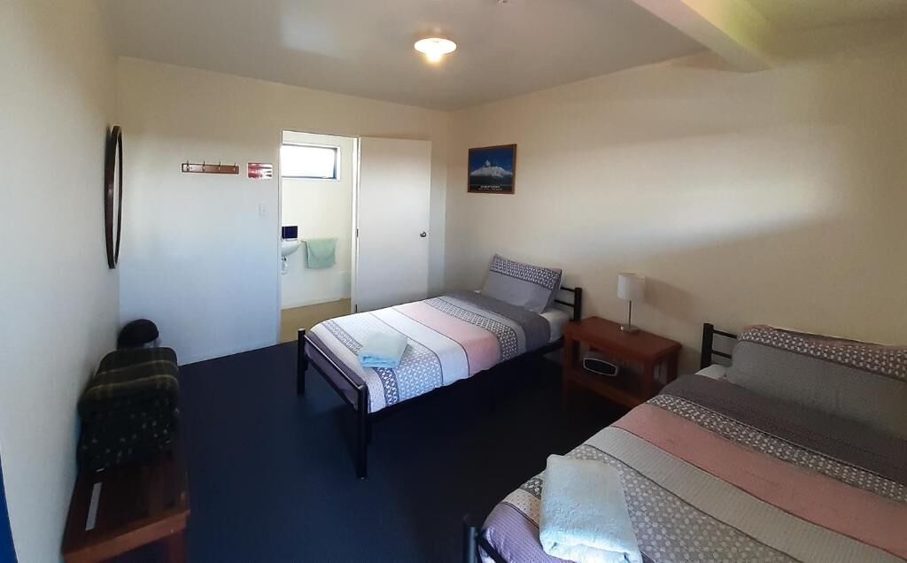 A room with two beds, a desk, a chair, a door, and a wall-mounted light fixture.