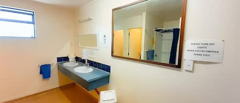 Un baño con dos lavamanos, un espejo, un cubo de basura y un cartel que indica apagar las luces.